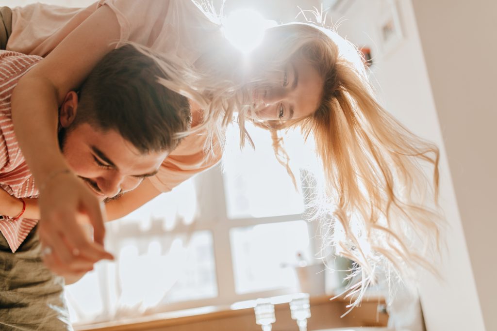 Positive blonde girl jumps on her lovers back. Portrait of young romantic couple fooling around in sunny room