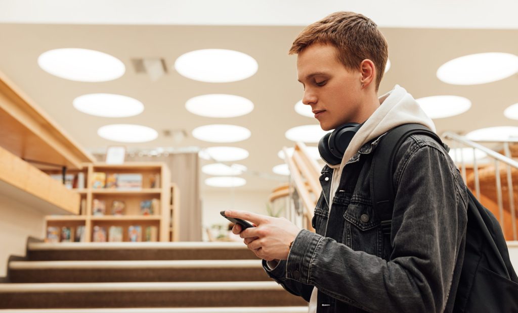 Young male with backpack and smartphone standing in library